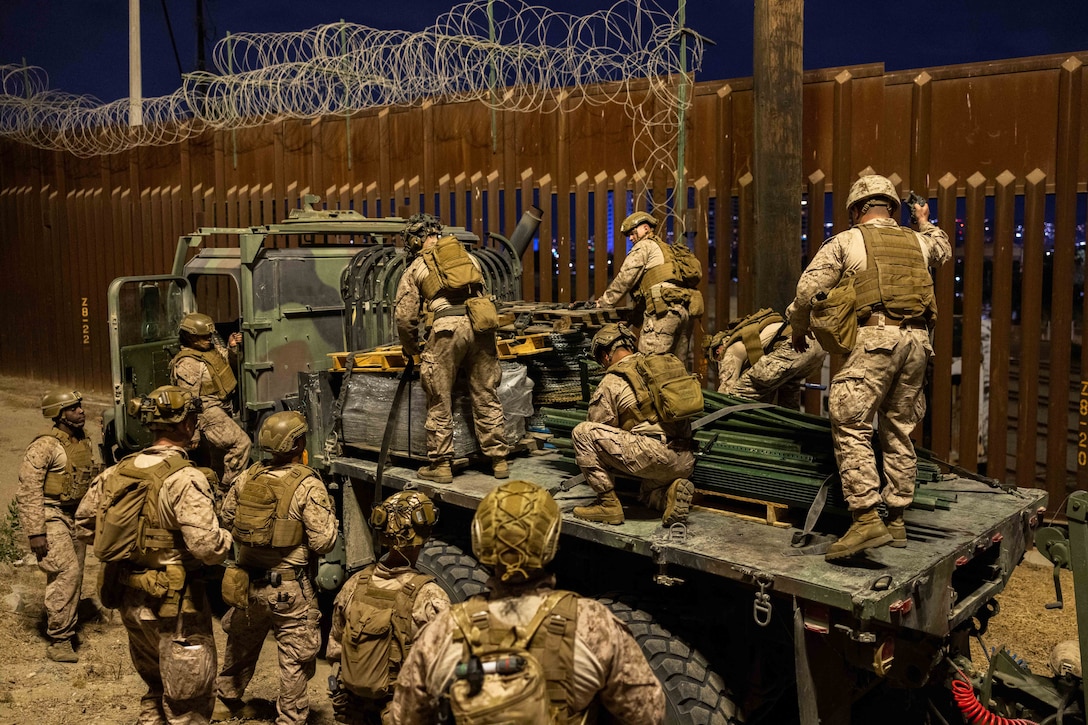 A group of Marines wearing uniforms stand on a flatbed truck to place wire along the top of a wall on the southern border of the U.S. while another group of Marines in uniform watch from the ground.