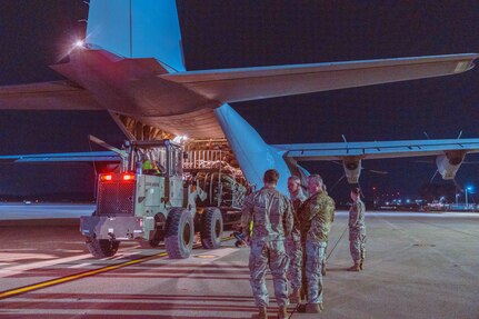 Three men in uniform stand near an aircraft while another man watches a truck unload the aircraft.
