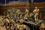 A group of Marines wearing uniforms stand on a flatbed truck to place wire along the top of a wall on the southern border of the U.S. while another group of Marines in uniform watch from the ground.