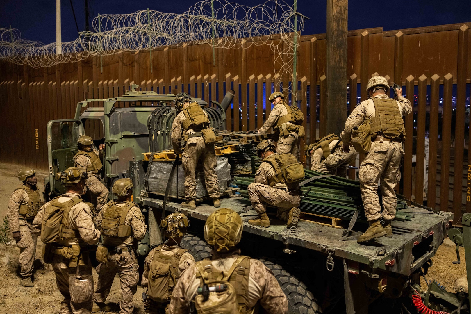 A group of Marines wearing uniforms stand on a flatbed truck to place wire along the top of a wall on the southern border of the U.S. while another group of Marines in uniform watch from the ground.