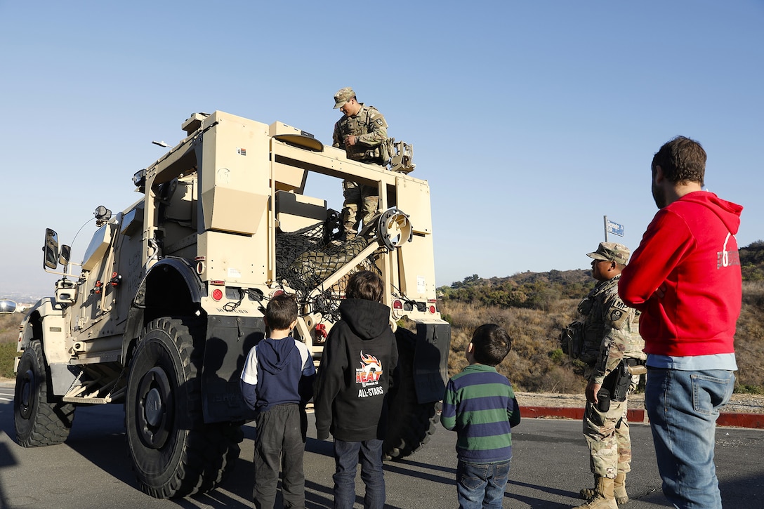 A soldier stands in the back of a military vehicle and talks to kids looking up at it from the street, where another soldier stands by.