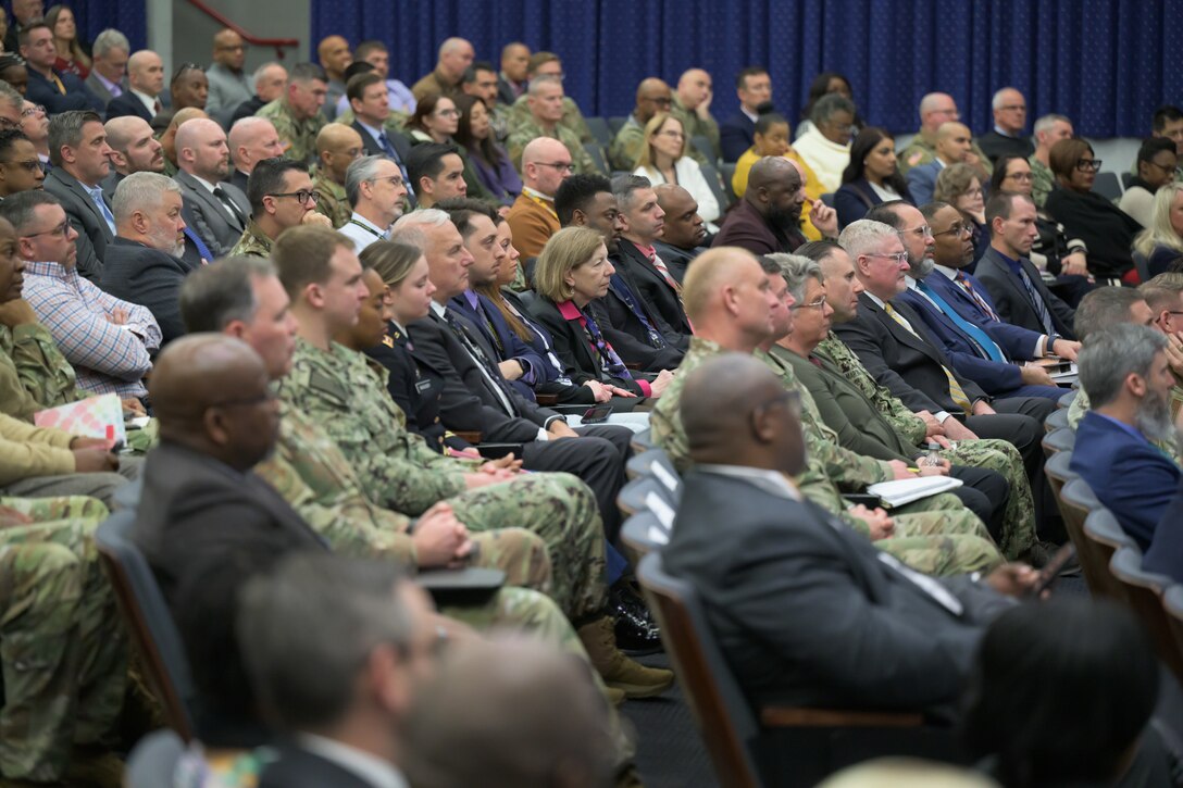 Men and women, some in military uniform, sit in an auditorium facing a stage