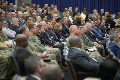 Men and women, some in military uniform, sit in an auditorium facing a stage