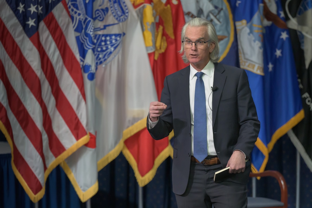 A white man stands on a stage in front of flags speaking