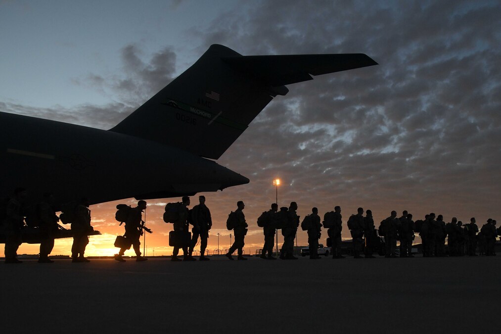 Dozens of soldiers carrying gear stand in a long line outside of a partially visible aircraft under a cloudy sky and sunlit peeking from the left.