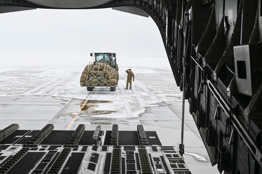 An airman stands on a snowy tarmac next to a forklift carrying supplies as seen from the open doorway of an aircraft.