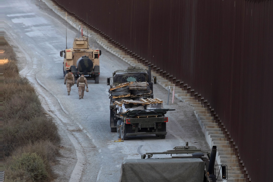 Two Marines walk beside two military vehicles on a winding road beside a tall metal fence.
