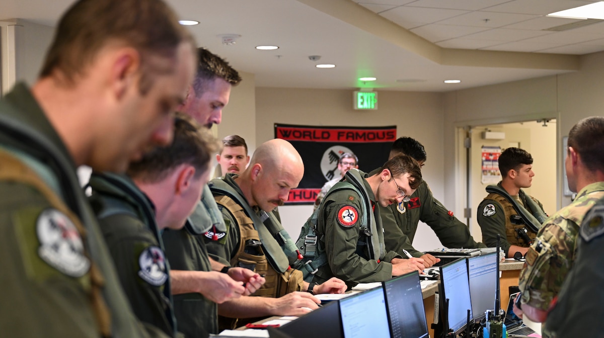 Ten pilots crowd into a briefing room and look over documents.