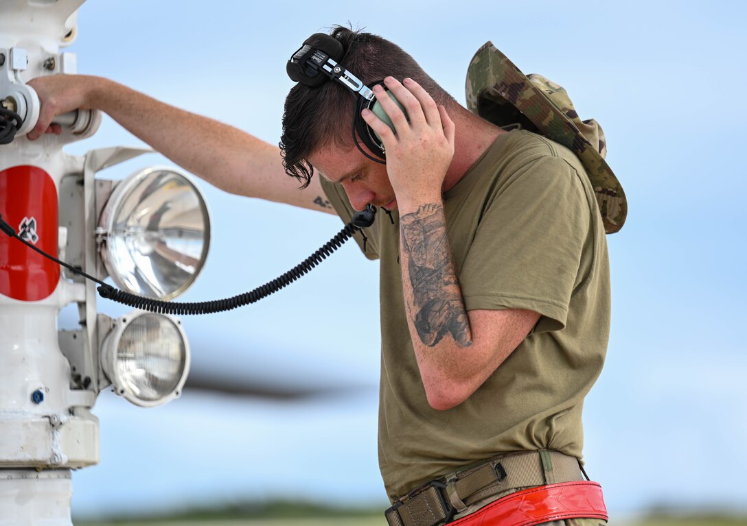 A man plugs his headphones into a port on the landing gear of a military aircraft and presses it into his ear with one hand.