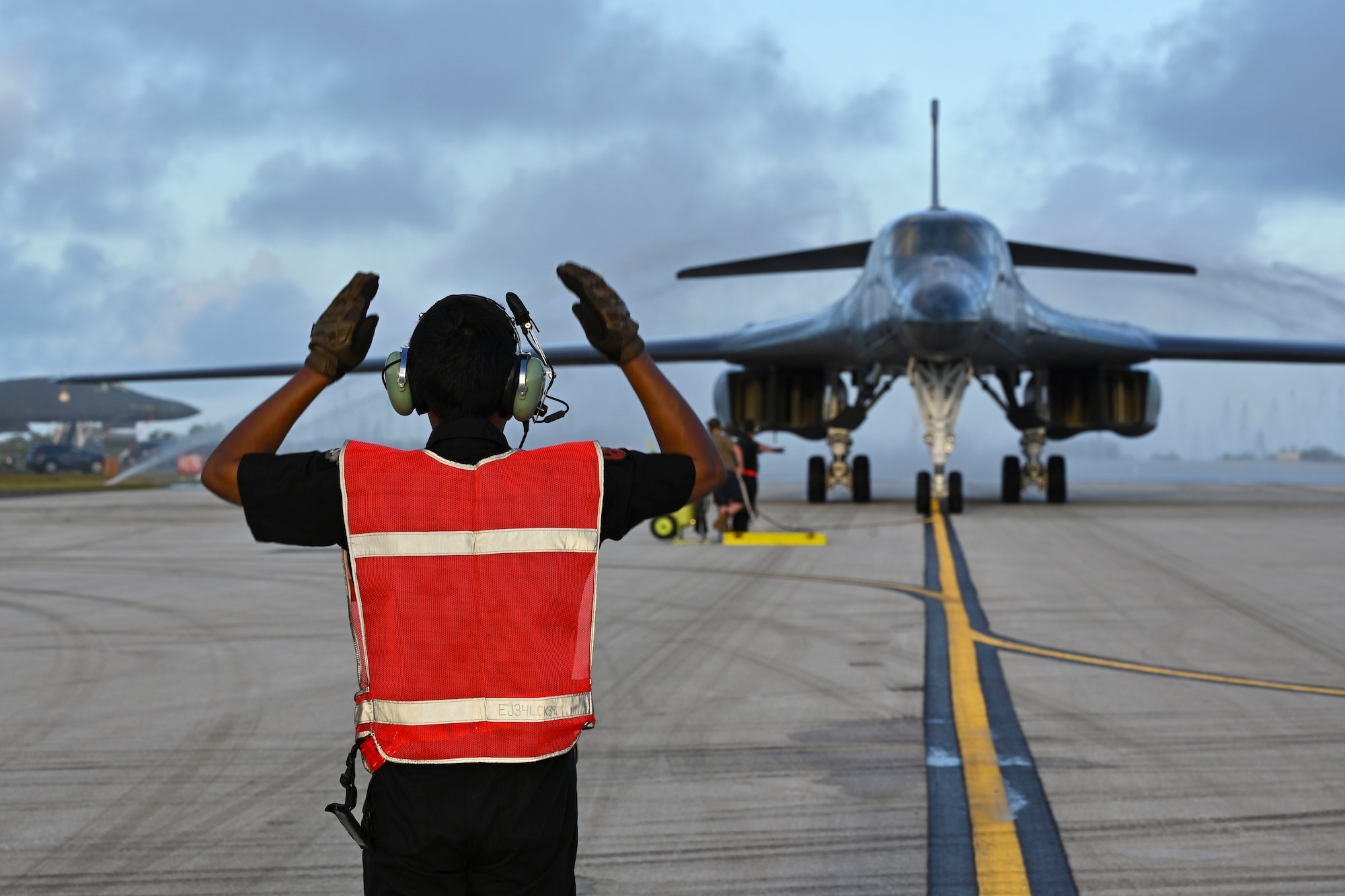 A man in a safety vest and headphone holds his hands in the air as a military jet rolls up towards him.