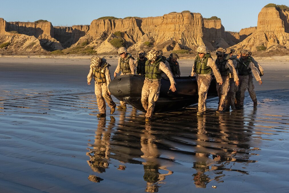 About a dozen Marines carry an inflatable boat through ankle-deep water with cliffs in the background.