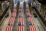 Sixteen service members in rows of four stand in formation aboard an aircraft with caskets covered in American flags between them.