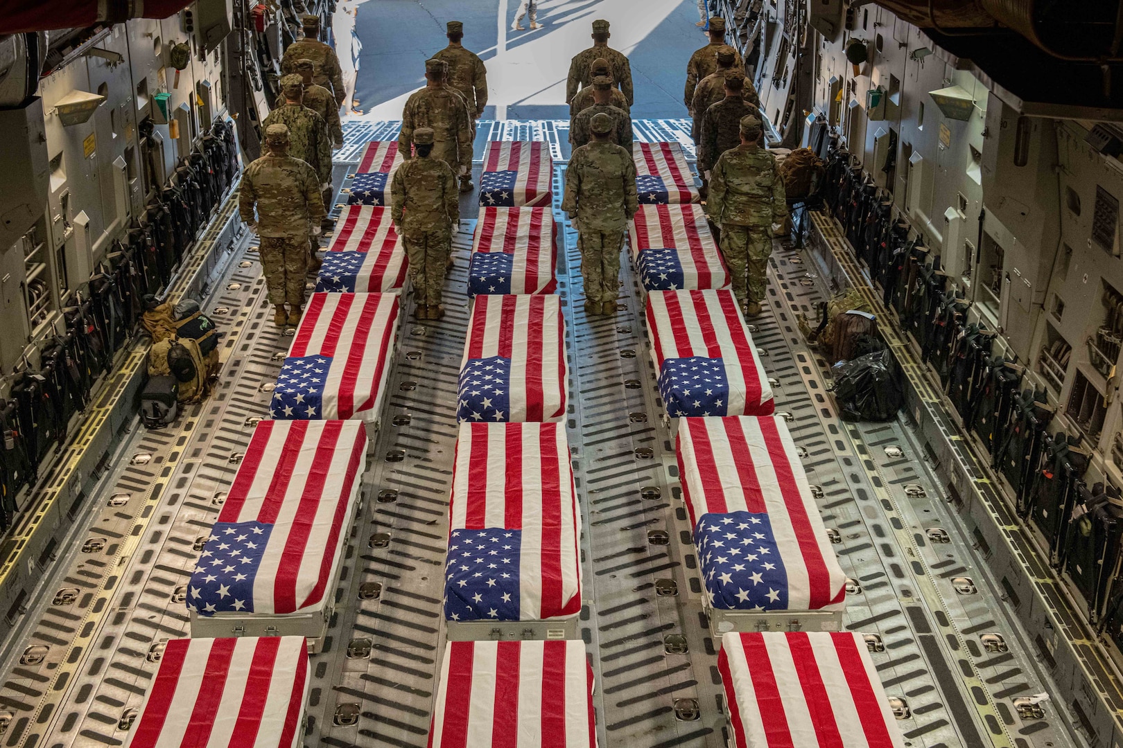 Sixteen service members in rows of four stand in formation aboard an aircraft with caskets covered in American flags between them.