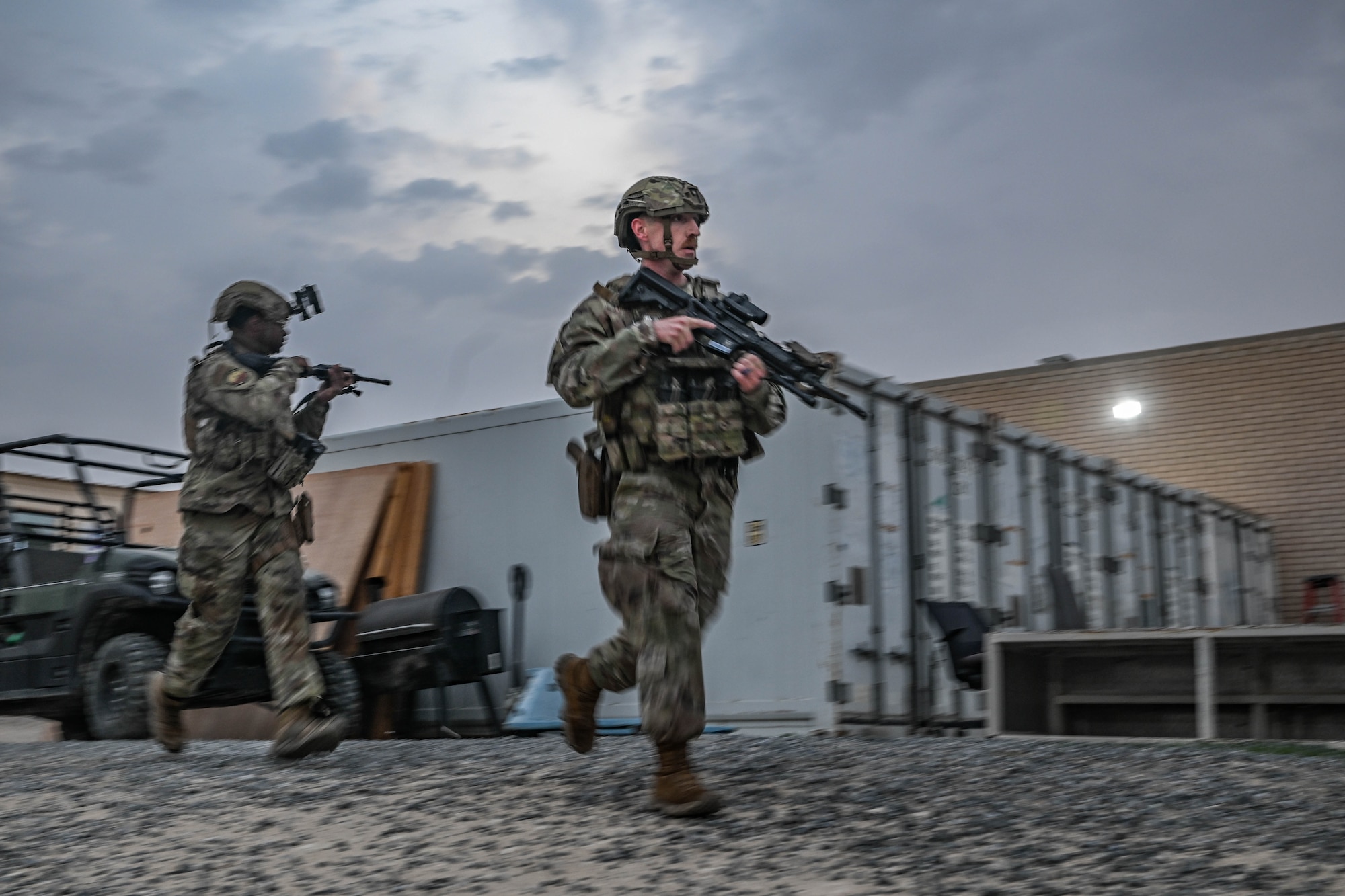 Two men in uniform carrying weapons sprint across a gravel path.