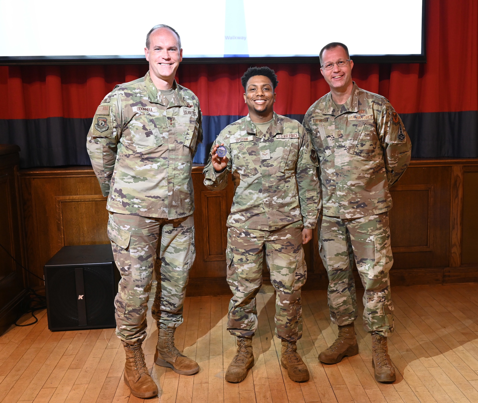 U.S. Air Force Maj. Gen. Jefferson O’Donnell, left, Air Force Personnel Center commander, Joint Base San Antonio-Randolph, Texas, coins Airman 1st Class Tyreq Upshaw, center, 100th Force Support Squadron Post Office postal clerk, for superior performance before hosting a town hall meeting with Chief Master Sgt. Edwin Ludwigsen, AFPC command chief, JBSA-Randolph, at RAF Mildenhall, England, Jan. 23, 2025. O’Donnell met with base leadership to increase awareness of talent management issues, enable better teamwork and execution, and receive feedback on how AFPC can better assist Airmen, Guardians and their families. (U.S. Air Force photo by Karen Abeyasekere)
