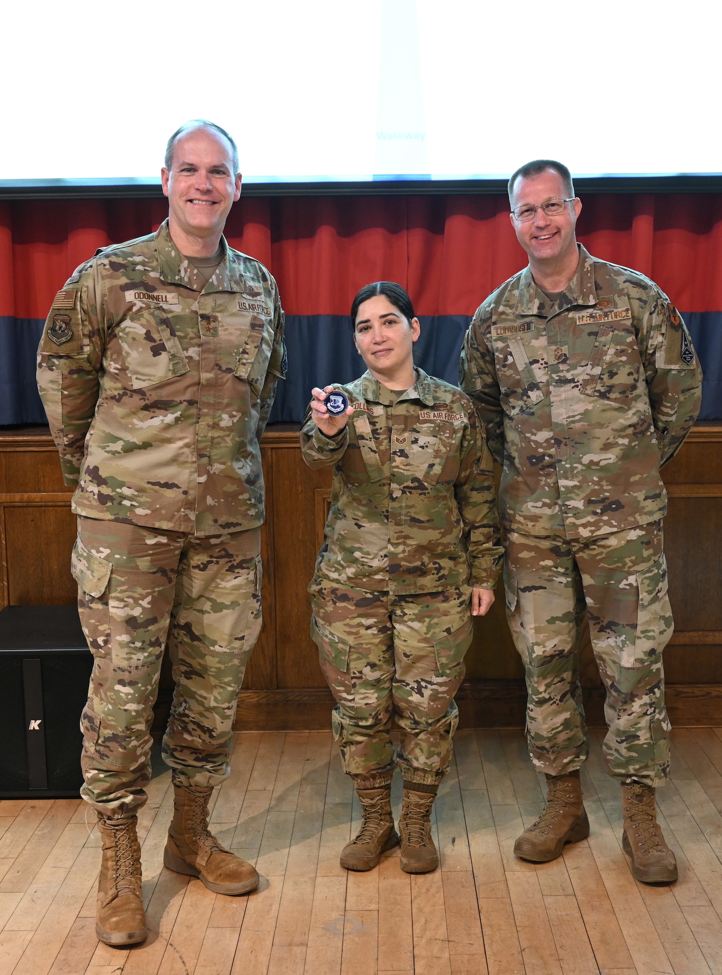 U.S. Air Force Maj. Gen. Jefferson O’Donnell, left, Air Force Personnel Center commander, Joint Base San Antonio-Randolph, Texas, coins Staff Sgt. Erin Collins, center, 100th Maintenance Squadron commander’s support staff, for superior performance before hosting a town hall meeting with Chief Master Sgt. Edwin Ludwigsen, AFPC command chief, JBSA-Randolph, at RAF Mildenhall, England, Jan. 23, 2025. O’Donnell met with base leadership to increase awareness of talent management issues, enable better teamwork and execution, and receive feedback on how AFPC can better assist Airmen, Guardians and their families. (U.S. Air Force photo by Karen Abeyasekere)