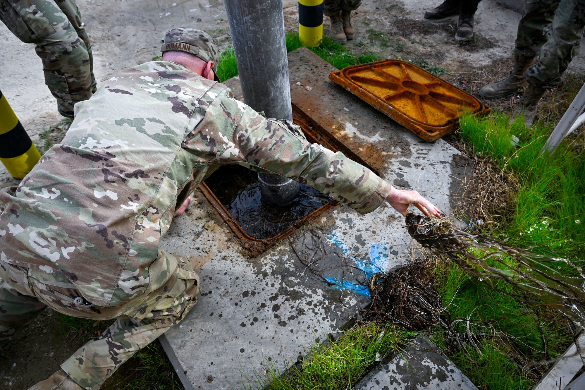 A U.S. Air Force Airman cleans out weed over growth in water while a hydro-excavation machine is in use.