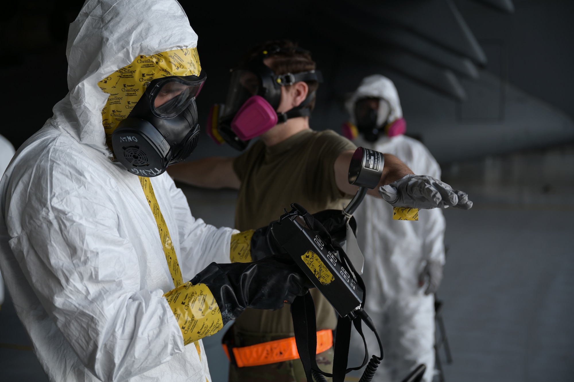An Airman dressed in chemical protection gear uses chemical detection equipment on an Airman not in protective gear.