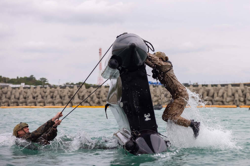 A Marine in water pulls a rope attached to a craft to flip it over while another Marine hangs on to the other side of the craft as it is flipped.