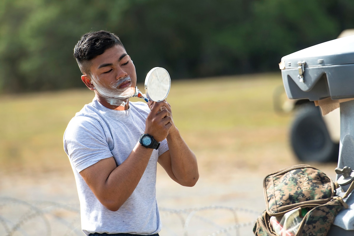 An airman holds up a circular mirror while shaving in a brown field with a backpack.
