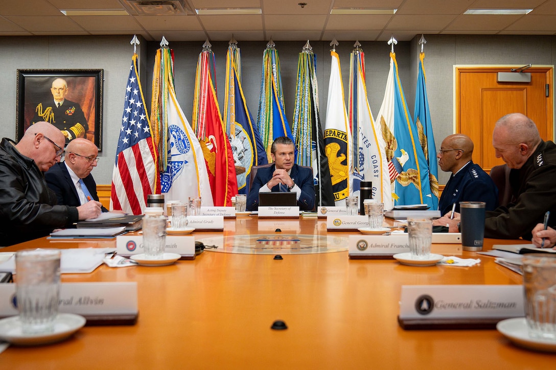 Defense Secretary Pete Hegseth sits at the head of a table with service leaders and flags standing behind him.
