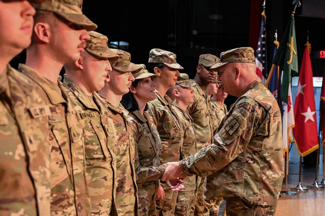 People in uniform stand on a stage as a man in uniform shakes their hand.