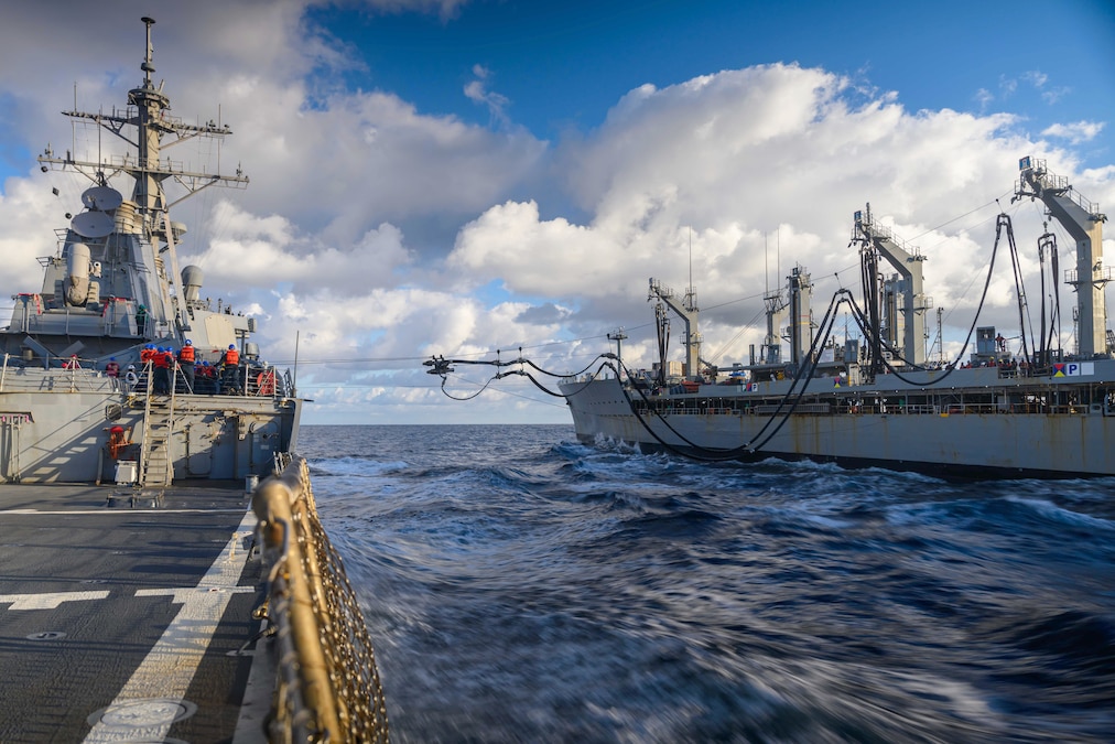 A black hose extends from one ship to another in dark waters under a cloudy, blue sky.