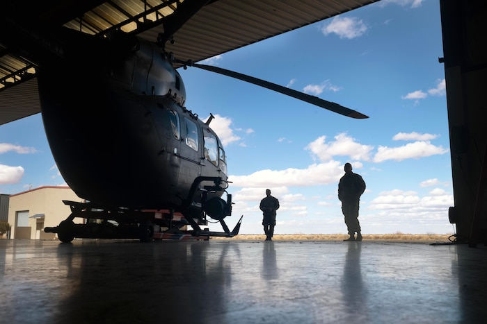 Two soldiers in silhouette look at a helicopter in a hangar bay while standing in a field under a cloudy, blue sky.