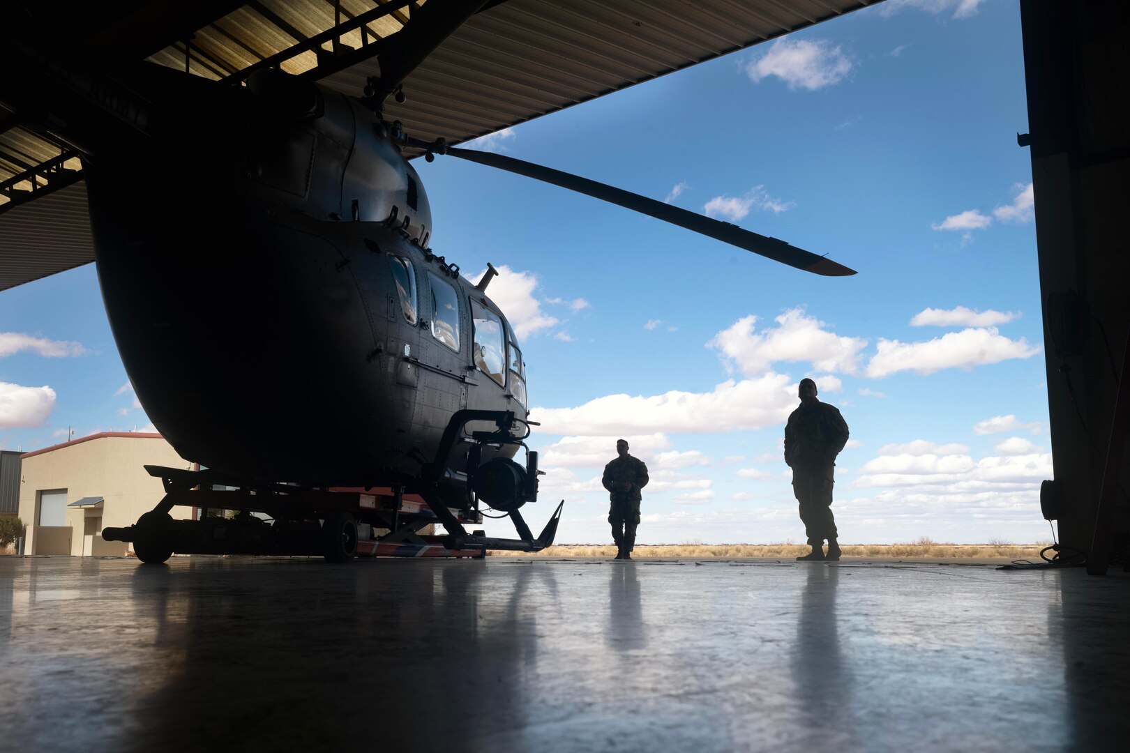 Two soldiers in silhouette look at a helicopter in a hangar bay while standing in a field under a cloudy, blue sky.