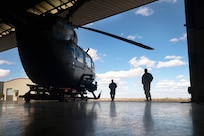 Two soldiers in silhouette look at a helicopter in a hangar bay while standing in a field under a cloudy, blue sky.