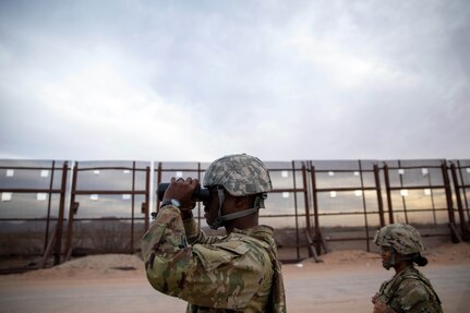 A soldier looks through binoculars as another soldier stands behind him. A metal fence is in the background.