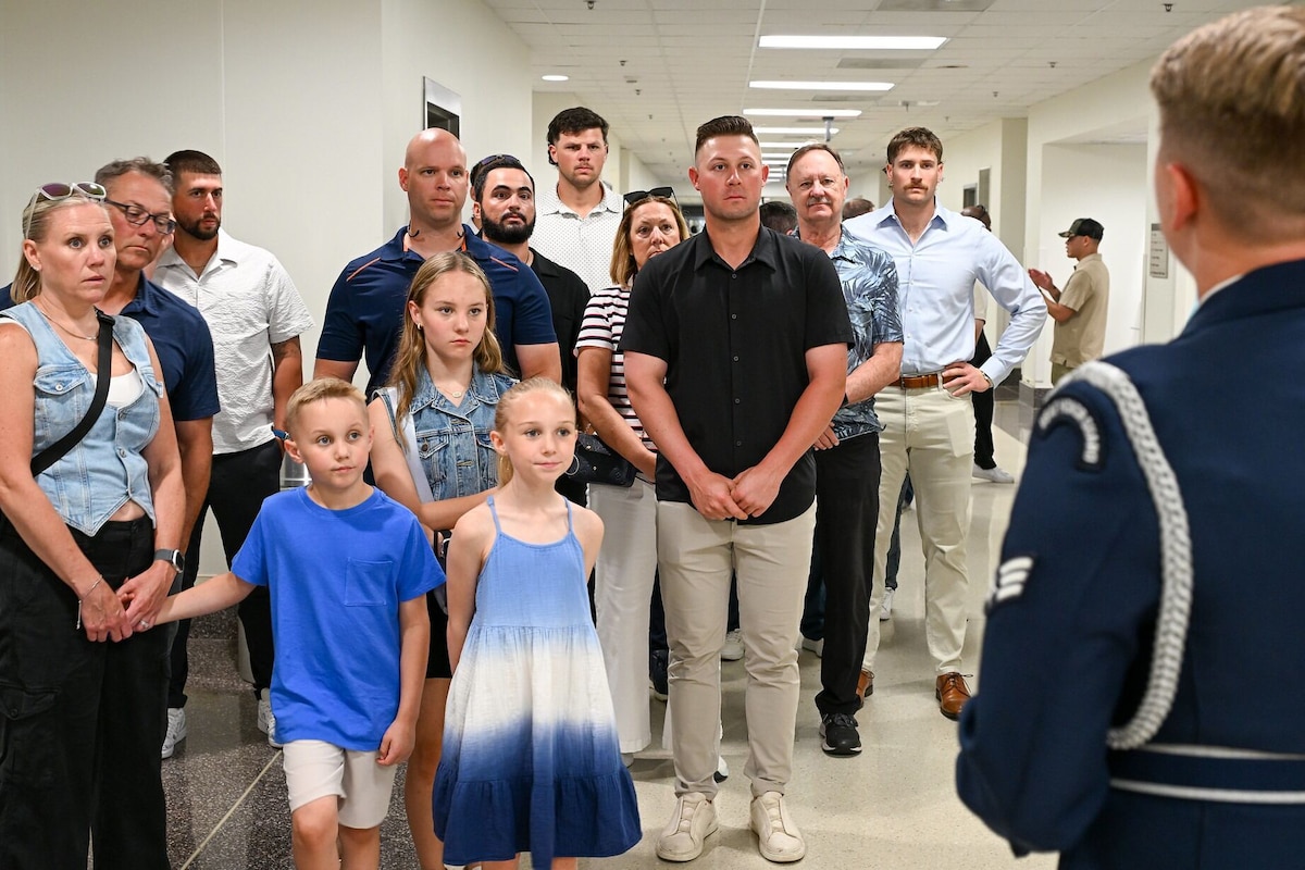 Men, women and children in summer casual clothes stand in a large hallway while listening to a man wearing a military dress uniform as he addresses the group in the foreground of the photo, his back to the camera.