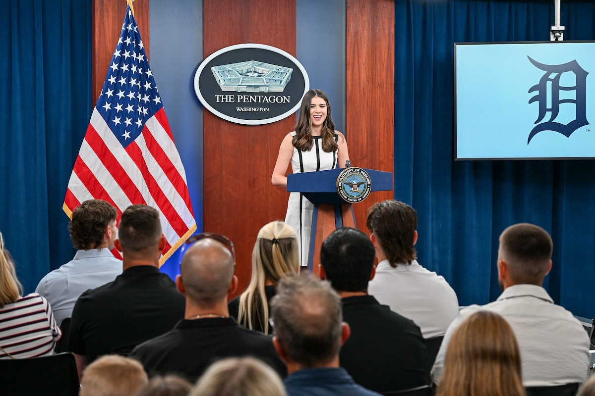A woman wearing a dress stands behind a podium on a stage, smiling as she speaks to a group of people sitting in front of her, their backs to the camera. An American flag is to her left, and behind her is a sign on the wall that shows a five-sided building and reads “The Pentagon – Washington.” The letter “D” is displayed on a screen to the right of the woman.
