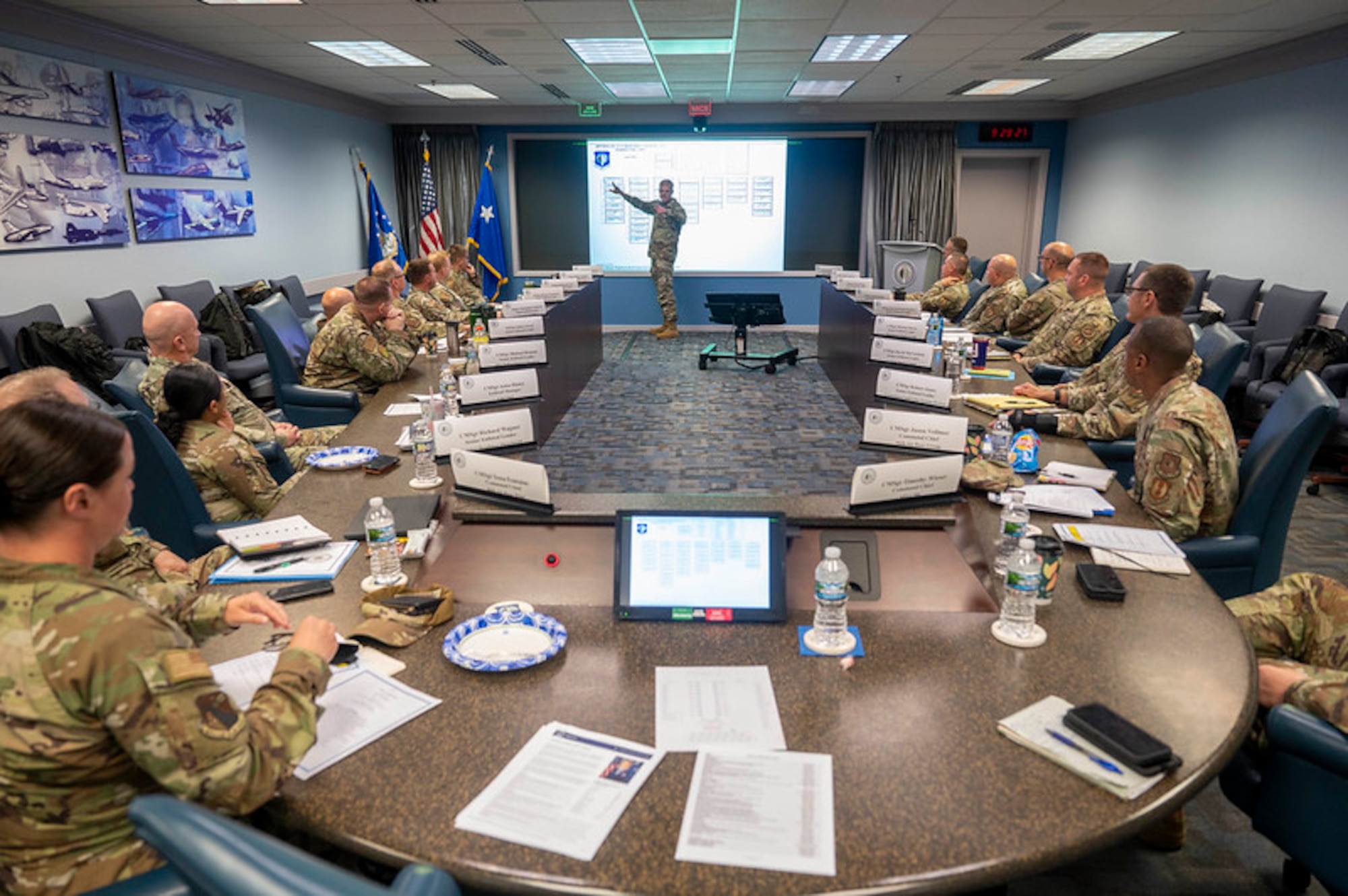 Air Force members watch man give presentation at big screen. Everyone is wearing fatigues.