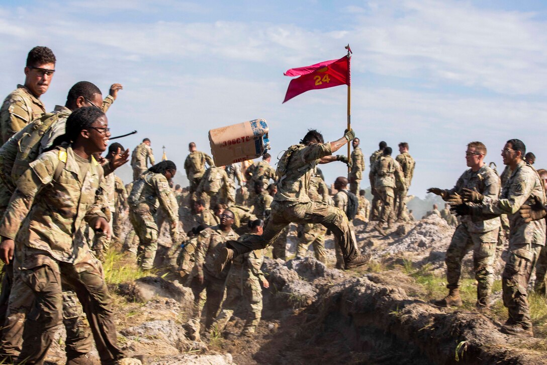 A soldier holding a red flag with a pole leaps over a trench as soldiers on one side prepare to jump and soldiers on the other side prepare to help.