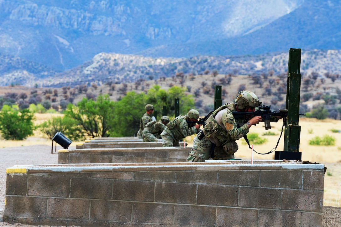 Four soldiers are seen at an outdoor gun range, three of them firing rifles; sparse vegetation and mountains are in the background.