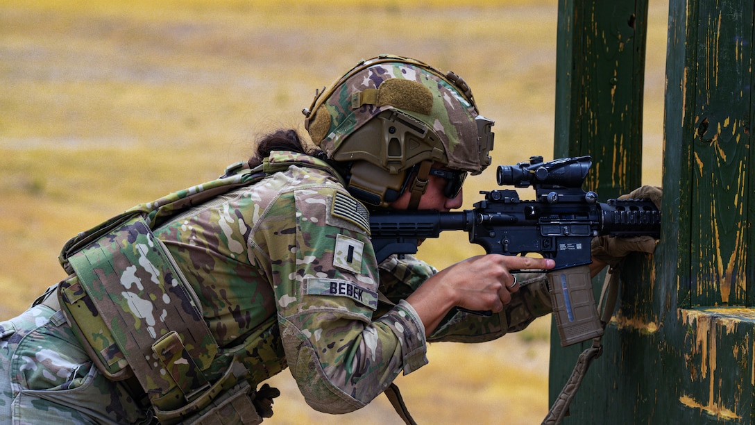 A soldier in combat gear steadies a rifle on a wooden frame outdoors and looks through the scope.