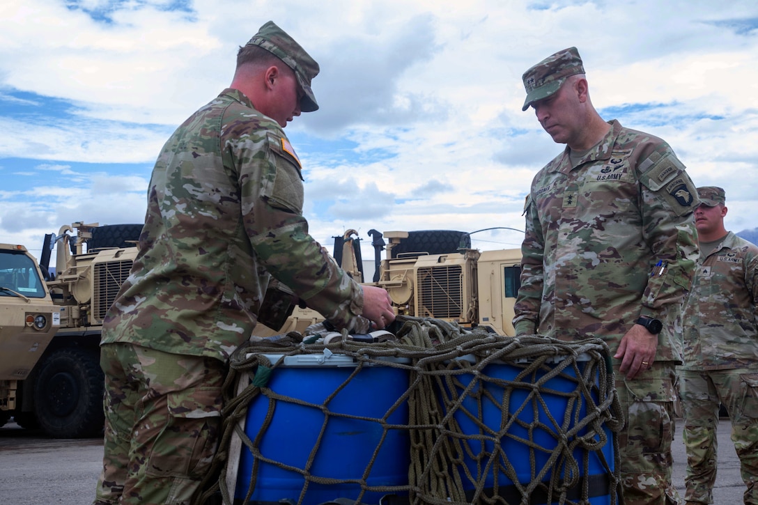 A soldier standing outdoors shows another how a large, net-like harness attaches to a blue container as a third soldier looks on.
