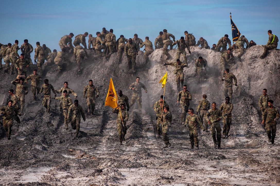 Soldiers climb over and run down a dirt berm; three carry poles with flags aloft.
