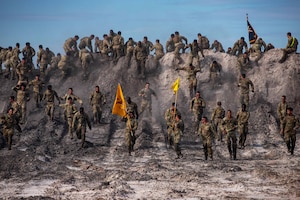 Soldiers climb over and run down a dirt berm; three carry poles with flags aloft.