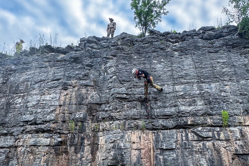 A soldier wearing a harness stands on a rocky wall while taking a picture as fellow soldiers stand near the edge of the cliff to watch.