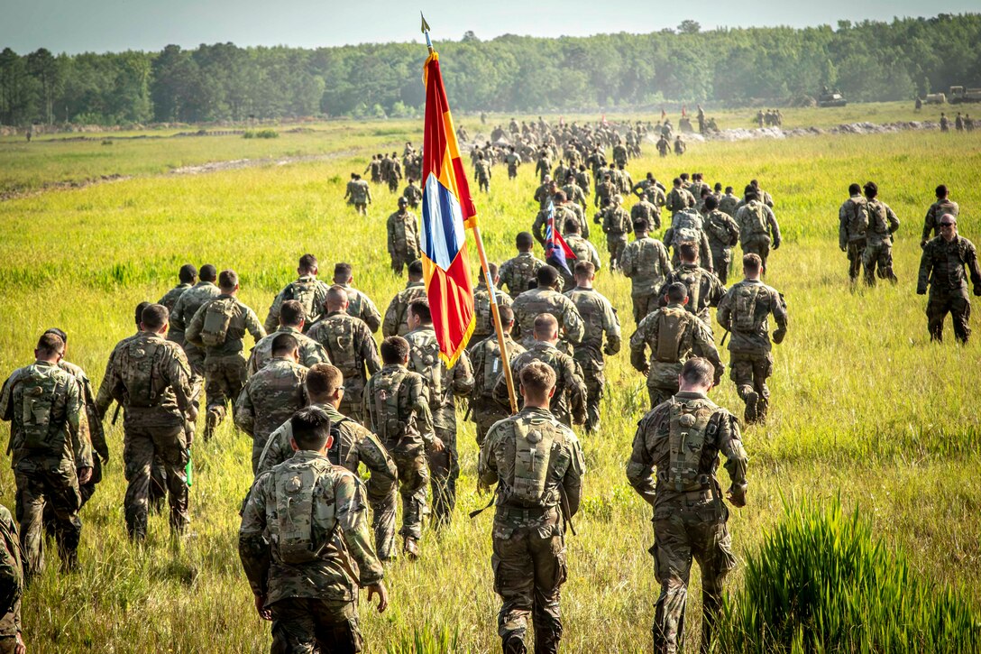 Soldiers walk or jog across a grassy field toward a line of trees in the distance; one holds a pole with a flag aloft.