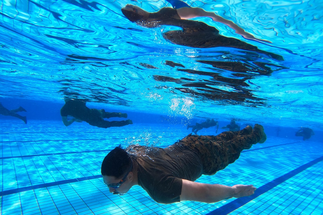 A Marine in uniform swims near the bottom of a pool as their reflection is seen at the surface and fellow swimmers are in the background.