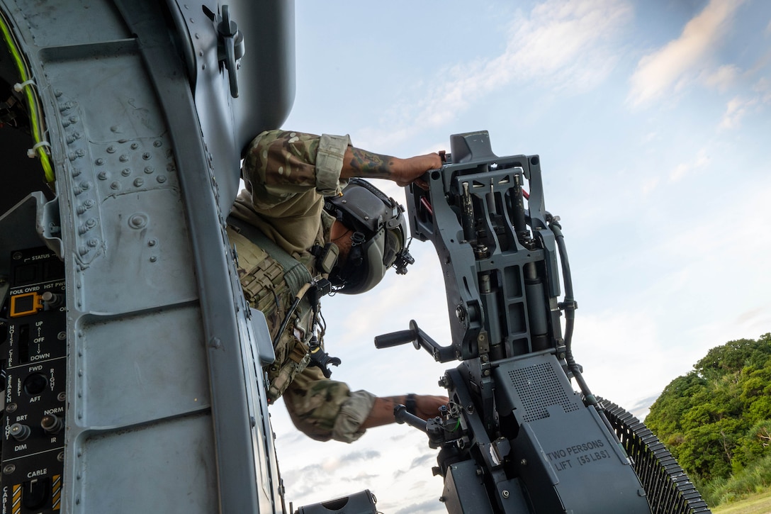An airman wearing protective gear leans out of a helicopter to fire a weapon under a blue sky.