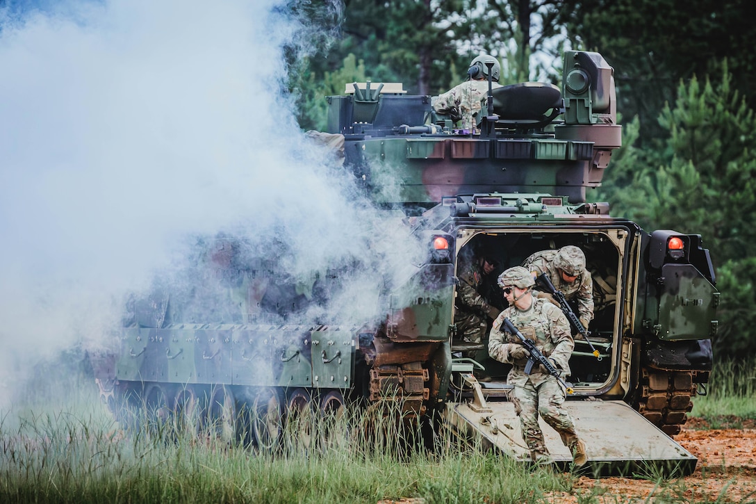 Smoke surrounds a camouflage tank as soldiers with weapons exit from the rear into a wooded area.