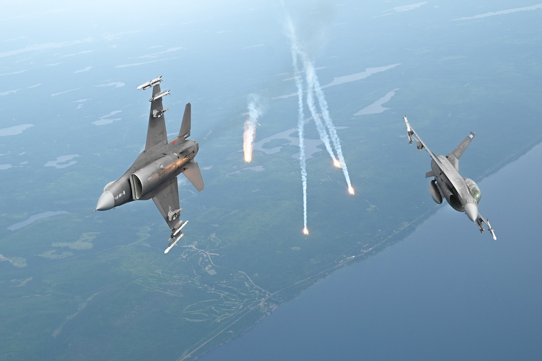 Two aircraft fly on their side as flares fly in between them in a hazy sky over a coastal area.