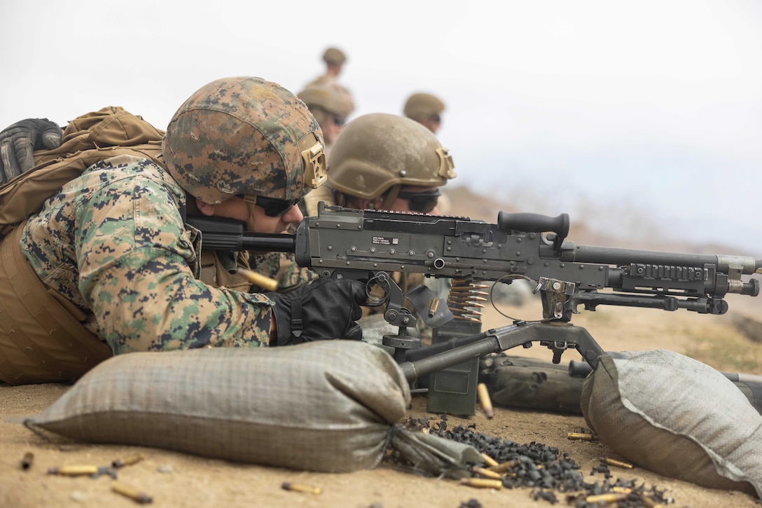 Ammo sits on a dirt ground as a Marine lies on sandbags while firing a weapon next to fellow Marines.