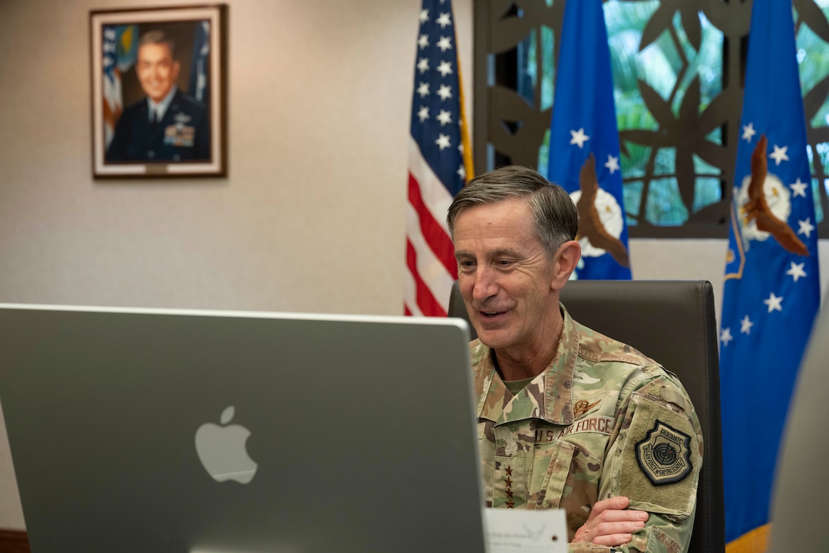A older man in uniform sits behind a computer screen with paintings of former commanders on the wall behind them.