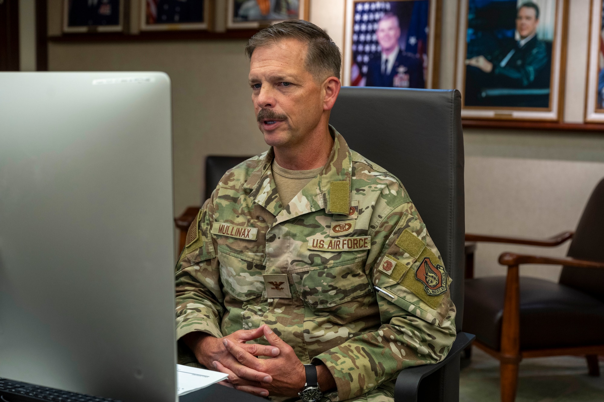 A older man in uniform sits behind a computer screen with paintings of former commanders on the wall behind them.