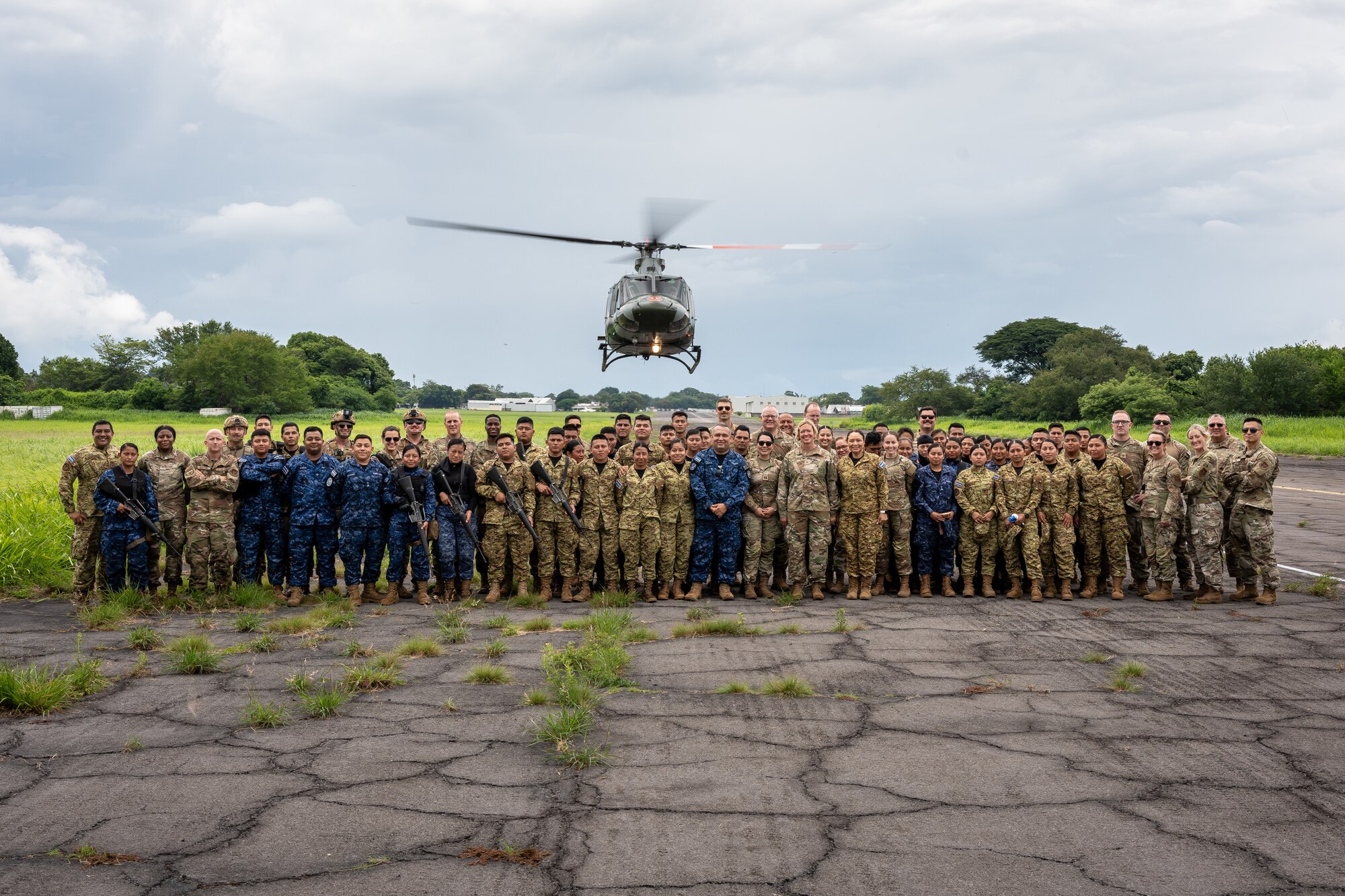 A group photo of international forces with a helicopter hovering in the background.